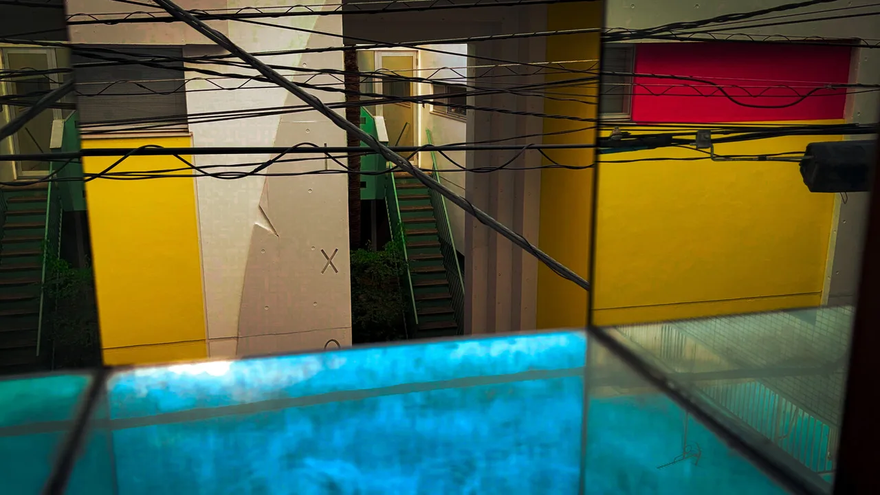 An apartment courtyard of yellow, white and red walls and green stairs, seen across a tangle of overhead wires and through panes of glass that glow turquoise below.