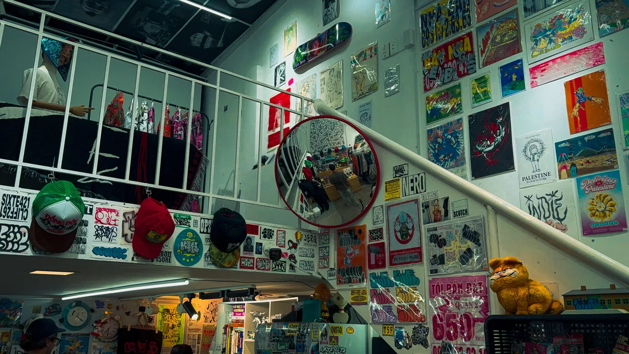 A small shop with every wall covered in posters and stickers, caps hanging from a stair rail and a round mirror reflecting the room.
