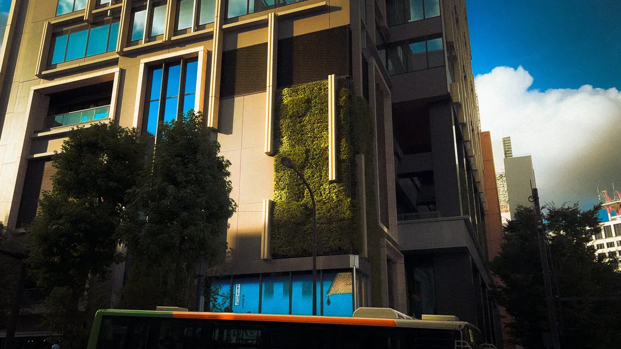 An office tower with a green living wall, a tree in front and a bus passing below a blue sky.