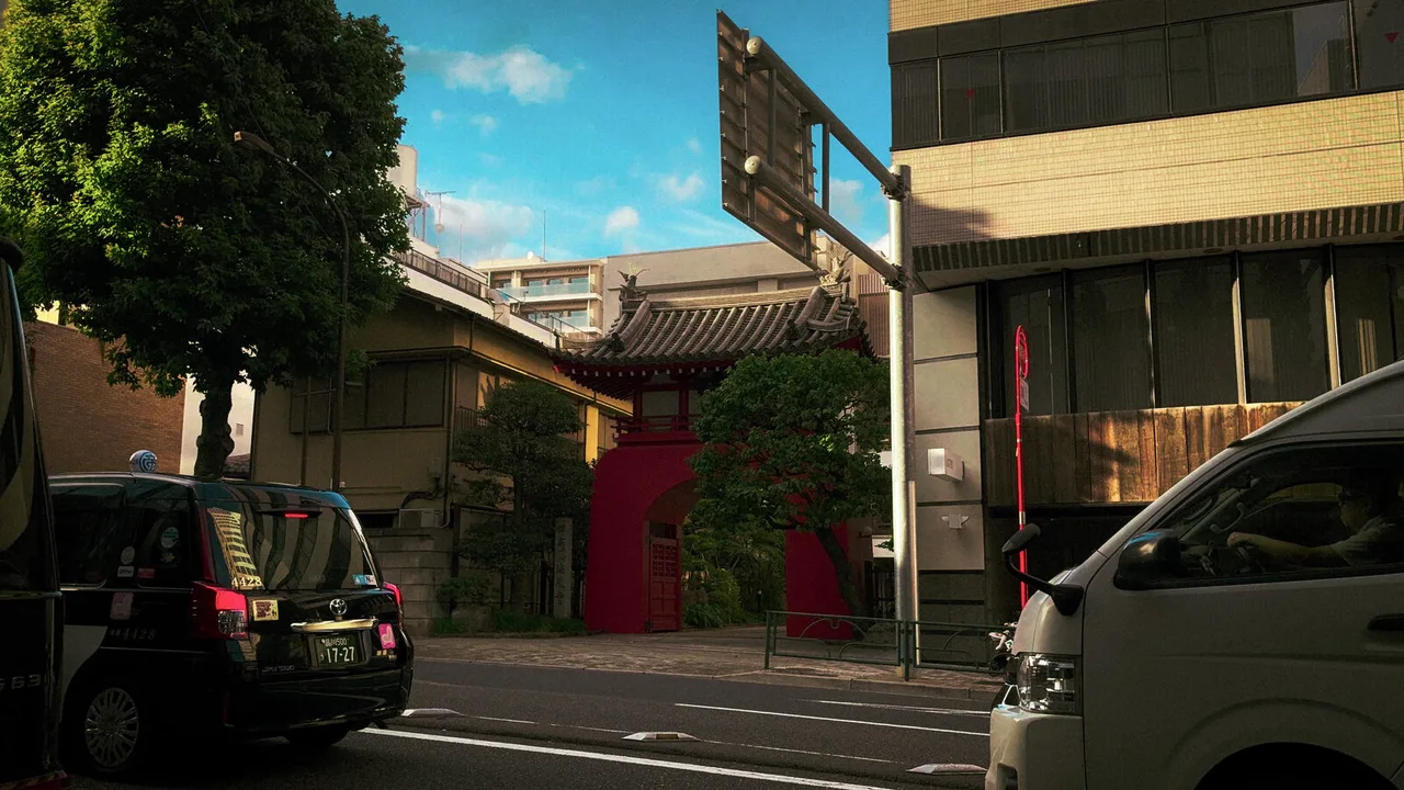 A quiet street on a bright day: a black taxi and a white van pass a red temple gate under a tiled roof.