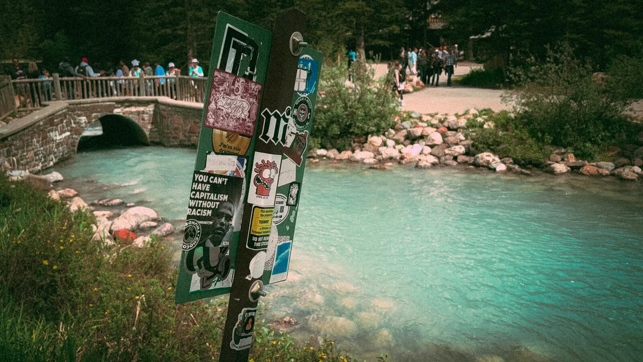 A sticker-covered signpost in the foreground; behind it a turquoise river, a small stone bridge and a crowd of visitors.