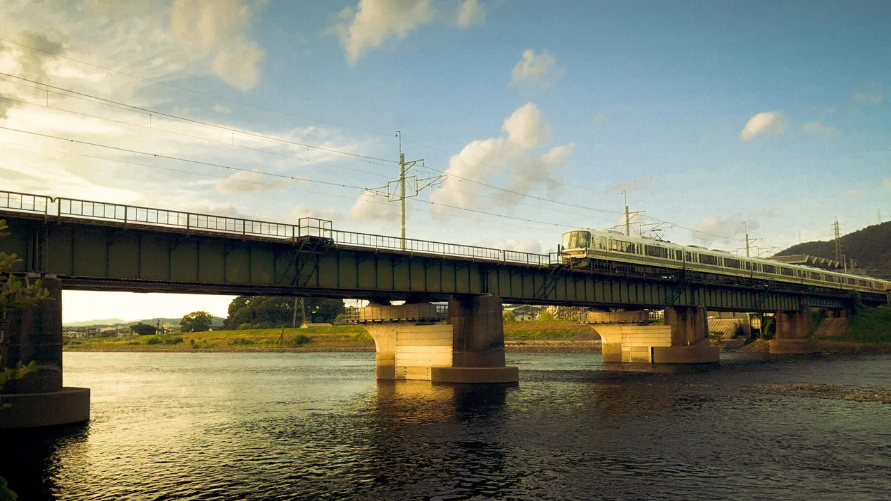 A green train crossing a long steel bridge over a river in low evening sun, clouds drifting overhead.
