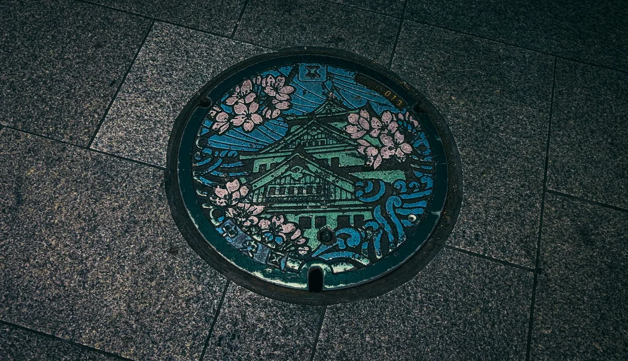 A painted manhole cover set in granite paving, showing a castle framed by pink cherry blossoms and waves.