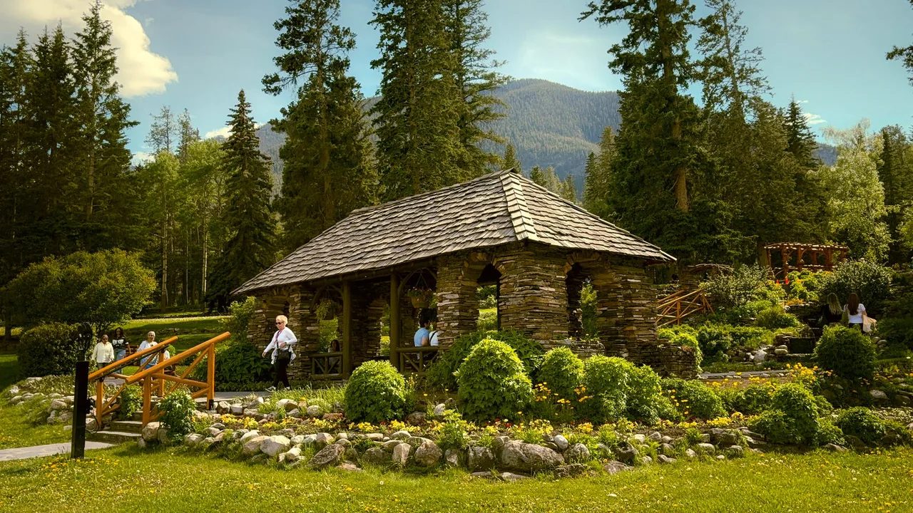 A stone and timber pavilion in a flower garden, tall pines around it and a mountain behind.