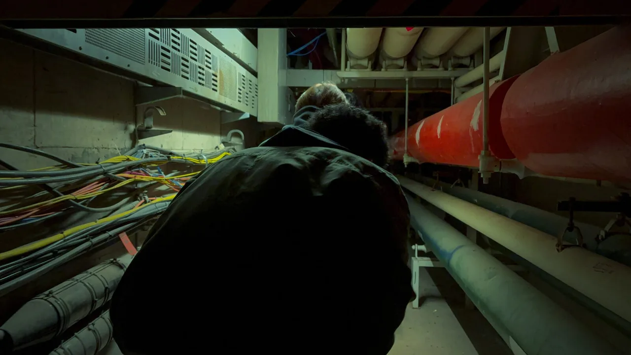 A figure seen from behind, stooping through a service tunnel packed with red and pale pipes and loose bundles of cable.