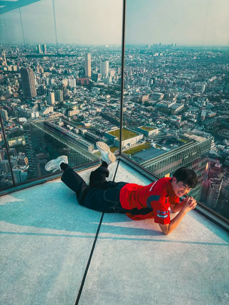 Donald Phan lying in the glass corner of a rooftop observation deck, chin on clasped hands, grinning, with a city spread out far below.